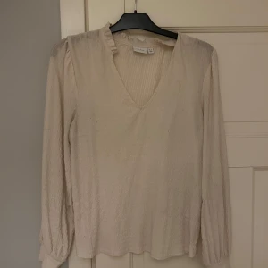 Beige blus från VILA - Säljer en elegant beige blus från VILA med v-ringning och långa ärmar. Blusen har en lätt textur och är perfekt för en stilren look. Passar bra till både jeans och kjol.