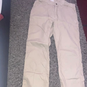Beige byxor från Carhartt - Säljer ett par beige byxor från Carhartt i 100% bomull. De har en relaxed fit och är perfekta för en avslappnad stil. Byxorna har en klassisk design med knappar och dragkedja.