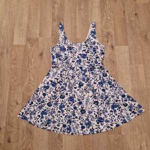 Blommig klänning med blått mönster - Säljer en söt klänning med blommigt mönster i blått och vitt. Klänningen har en kort längd och är ärmlös, perfekt för varma dagar. Den har en rund halsringning och en lätt utställd kjol som ger en fin silhuett.