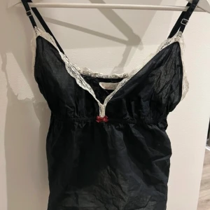 Svart linne med spetsdetaljer - Säljer ett svart linne från Odd Molly med vita spetsdetaljer och en liten röd rosett framtill. Linnet har smala axelband och en dekorativ logga på baksidan. Perfekt för en unik och stilren look.