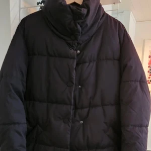 Svart dunjacka från Monki - Säljer en svart dunjacka från Monki i storlek XS. Jackan har en hög krage och knäppning framtill. Perfekt för kalla dagar med sin långa passform och vadderade design. En stilren och praktisk jacka för vintern. 
