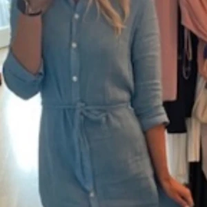 Lexington- Ljusblå klänning med knytband - Ny, endast prövad. Säljer en stilren ljusblå klänning från Lexington  med knytband i midjan och knappar framtill. Klänningen har långa ärmar och en avslappnad passform, perfekt för en casual look. Passar bra till både vardag och lite finare tillfällen.