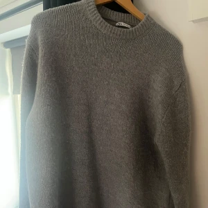 Grå stickad tröja från Zara - Säljer en stilren grå stickad tröja från Zara. Perfekt för kyligare dagar med sin mjuka och bekväma passform. Tröjan har långa ärmar och en rund halsringning. Ett måste i garderoben för en klassisk look.