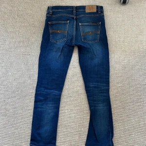 Blå jeans från Nudie Jeans Grim Tim 28W 32L - Snygga blå jeans från Nudie Jeans med klassisk femficksdesign och orange sömmar. De har en dragkedja och knapp framtill samt märkets logga på baksidan. Perfekta för en stilren och grich look.