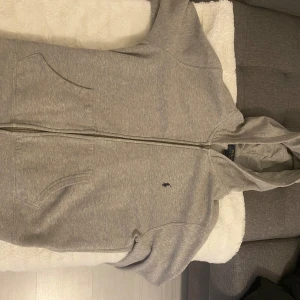 Grå hoodie från Ralph Lauren - Säljer en stilren grå hoodie från Ralph Lauren med dragkedja och fickor framtill. Perfekt för en avslappnad look. Den har en liten broderad logga på bröstet och är tillverkad i mjukt material.