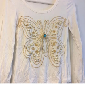 Vit långärmad tröja med fjärilsbroderi storlek XS/S - Säljer en vit långärmad tröja med ett stort fjärilsbroderi på framsidan. Fjärilen är dekorerad med pärlor och en blå sten i mitten. Perfekt för att ge din outfit en unik touch!