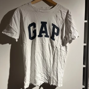 Vit t-shirt från GAP - Säljer en klassisk vit t-shirt från GAP med deras ikoniska logga i mörkblått på framsidan. T-shirten är kortärmad och tillverkad i mjuk bomull, perfekt för en avslappnad stil.
