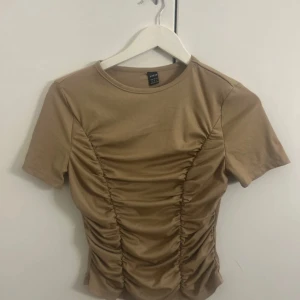 Beige ribbad topp från Shein - Säljer en beige ribbad topp från Shein med korta ärmar och snygg rynkdetalj framtill. Perfekt för en stilren look.