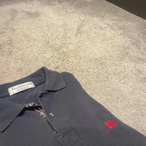 Mörkblå pikétröja från Burberry - Säljer en stilren mörkblå pikétröja från Burberry med klassisk krage och knappar framtill. Tröjan har en röd broderad logotyp på bröstet och är perfekt för en avslappnad men snygg look.