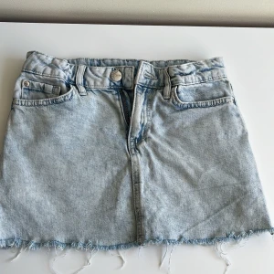 Ljusblå jeanskjol - Trendig ljusblå jeanskjol med rå kant och klassisk femficksdesign. Perfekt för en avslappnad stil. Dragkedja och knapp framtill för enkel stängning.