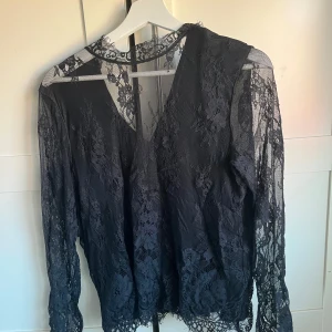 Svart spetsblus - Elegant svart blus i spets med långärmade ärmar och v-ringad design. Perfekt för en stilfull look med fina spetsdetaljer.