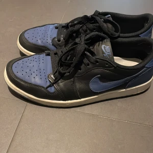 Nike Air Jordans i svart och blått - Snygga Nike Air Jordans i svart och blått med klassisk snörning. Skorna har en stilren design med perforerade detaljer på tån och en vit sula som kontrasterar mot det mörka skinnet.