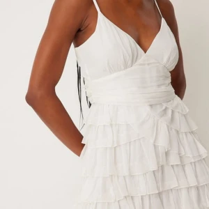 Vit klänning med volanger - Sperfin vit klänning med volanger från NAKD. Klänningen har en v-ringad design och snörning i ryggen för en perfekt passform. Perfekt för studenten💫