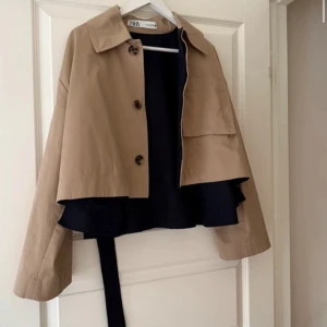 Beige och svart trenchcoat från ZARA - Snygg beige trenchcoat från ZARA med knappar framtill. Jackan har en klassisk krage och är croppad i modellen, vilket ger en modern touch. Perfekt för övergångsperioder!