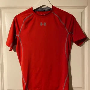 Röd kompressionströja från Under Armour - Säljer en röd kompressionströja från Under Armour med korta ärmar och gråa sömmar. Perfekt för träning med HeatGear-teknologi som håller dig sval och torr. Logotypen syns både fram och bak. Passar utmärkt för intensiva träningspass.