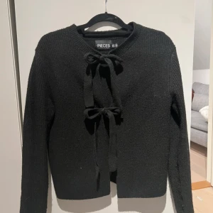 Svart stickad kofta från Pieces - Säljer en svart stickad kofta från Pieces med knytband framtill. Koftan har långa ärmar och en enkel design som passar perfekt för kyligare dagar. Perfekt att ha över en topp eller klänning för extra värme. Står xxs men sitter som xs/s 🫶