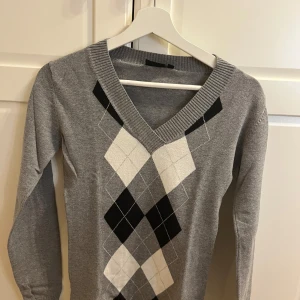 Grå tröja med argylemönster från Equipe - Snygg grå tröja från Equipe med klassiskt argylemönster i svart och vitt. Tröjan har en v-ringad hals och långa ärmar, perfekt för en stilren look. Passar bra till både jeans och kjol.