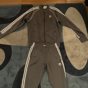 Grå träningsdräkt från Adidas - Säljer en grå träningsdräkt från Adidas med klassiska vita ränder längs ärmar och ben. Jackan har dragkedja och byxorna har elastisk midja. Perfekt för en sportig look eller avslappnad stil.