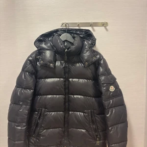Svart dunjacka från Moncler - Snygg svart dunjacka från Moncler med huva och dragkedja. Jackan har långa ärmar och en logotyp på ärmen. Perfekt för kalla dagar med sin quiltade design. Den är i storlek 2 vilket motsvarar M ungefär.