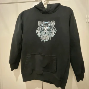 Svart hoodie från Kenzo - Säljer en svart hoodie från Kenzo med ett coolt tigertryck i blått och vitt på framsidan. Tröjan har en klassisk känguruficka och en bekväm huva. Perfekt för en avslappnad stil.