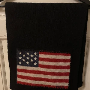 Svart halsduk från Polo Ralph Lauren - Snygg svart halsduk från Polo Ralph Lauren med en amerikansk flagga i rött, vitt och blått. Tillverkad i 100% ull, perfekt för att hålla dig varm under kyliga dagar. Flaggan ger en cool touch till en klassisk design.