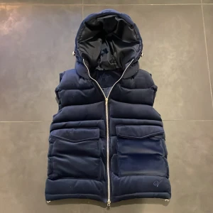 vendiera väst - Snygg mörkblå dunväst med dragkedja och stor huva. Västen har två stora fickor framtill och är perfekt för kyligare dagar. Den är både stilren och praktisk.