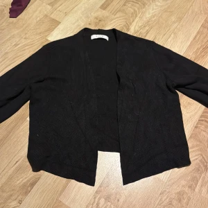 Svart ribbad kofta från VILA - Säljer en stilren svart ribbad, trekvarts kofta från VILA. Perfekt för lager-på-lager och har en öppen design utan knappar. Passar bra till både vardag och mer uppklädda tillfällen.