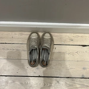 Beige sneakers från Axel Arigato - Säljer ett par stilrena beige sneakers från Axel Arigato. Skorna har en klassisk design med snörning och en diskret logga på hälen. Perfekta för en avslappnad look.