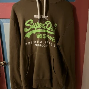 Brun hoodie från Superdry - Säljer en brun hoodie från Superdry med grön och vit text på framsidan. Den har en klassisk känguruficka och justerbar huva med vita snören. Perfekt för en avslappnad stil.