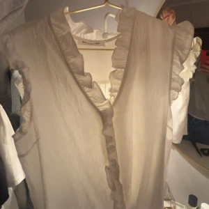 Vit/beige blus med volangdetaljer - Säljer en stilren beige blus med volangdetaljer längs ärmarna och en v-ringad hals. Perfekt för en elegant look.