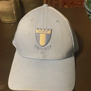 Ljusblå keps från Malmö FF - Säljer en ljusblå keps från Malmö FF med broderad logga framtill. Kepsen har justerbar rem baktill med MFF-broderi. Perfekt för fotbollsfans som vill visa sitt stöd.