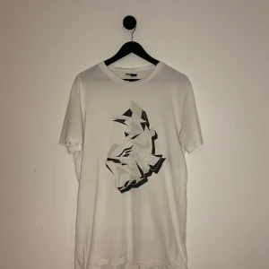 Lanvin T-shirt - Säljer en stilren vit t-shirt från Lanvin med ett coolt grafiskt tryck i svart och grått på framsidan. Perfekt för dig som gillar unika designer. T-shirten har en klassisk passform och korta ärmar. Storlek L men kan passa M 