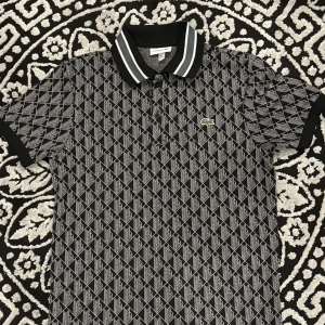 Lacoste pikétröja - Säljer en stilren svart och vit pikétröja från Lacoste med ett geometriskt mönster. Tröjan har korta ärmar och en klassisk krage med vita ränder. Perfekt för sommaren. Hör av er vid frågor 