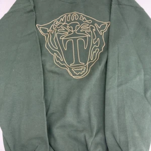 Grön tröja med tigertryck - Snygg grön tröja med ett stort tigertryck i beige på magen. Perfekt för en avslappnad stil med en unik touch. Tröjan har långa ärmar och en normal passform.