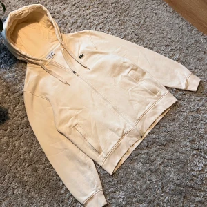 Beige hoodie från Lacoste - Säljer en stilren beige hoodie från Lacoste med dragkedja och den klassiska krokodilloggan på bröstet. Tröjan har en justerbar huva med snören och ribbade muddar vid ärmslut och nederkant. Perfekt för en avslappnad look.
