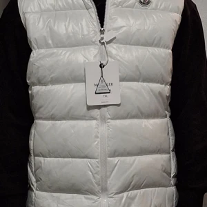 Vit dunväst från Moncler - Säljer en stilren vit dunväst från Moncler. Västen har en dragkedja framtill och är perfekt för kyligare dagar. Den är lätt och bekväm att bära, med Monclers logga på bröstet. Passar bra för både vardag och utomhusaktiviteter.