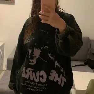 Cool svart oversized sweatshirt med tryck av The Cure på framsidan. Tröjan har en vintage-look med ett stort tryck i lila och vit text. Perfekt för fans av bandet eller för en avslappnad stil.