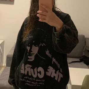 Svart sweatshirt med The Cure-tryck - Cool svart oversized sweatshirt med tryck av The Cure på framsidan. Tröjan har en vintage-look med ett stort tryck i lila och vit text. Perfekt för fans av bandet eller för en avslappnad stil.