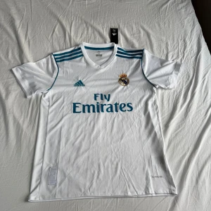 Vit Real Madrid fotbollströja från Adidas - Säljer en vit Real Madrid fotbollströja från Adidas med korta ärmar och klubbens emblem på bröstet. Tröjan har tre blå ränder på axlarna och Fly Emirates som sponsorlogga. Perfekt för alla fans som vill visa sitt stöd för laget!