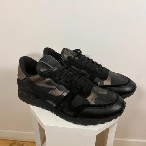 Rockrunners - Snygga Valentino Rockrunner sneakers i svart och grått med ett coolt camouflage-mönster. Skorna har snörning och är tillverkade i en kombination av mocka och syntetmaterial. Perfekta för en stilren look. Äkta såklart, skicket är som nya. Kan gå ned i pris vid snabb affär