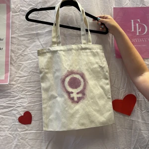 Vit shoppingväska med kvinnosymbol - Vi är ett UF företag som säljer tygväskor med handmålade tryck på framsidan. Perfekt för att bära med sig det nödvändigaste med stil.