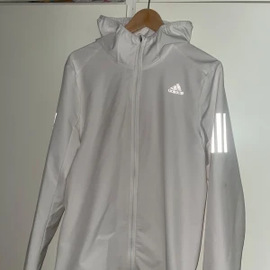 Vit vindjacka från Adidas - Säljer en stilren vit vindjacka från Adidas med reflekterande detaljer på ärmarna och logotypen på bröstet. Jackan har en dragkedja framtill och en praktisk huva. Perfekt för blåsiga dagar!