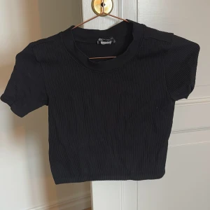 Svart ribbad topp - Säljer en stilren svart ribbad topp med korta ärmar. Perfekt för en avslappnad look. Toppen har en enkel design som gör den lätt att matcha med olika outfits. Liten i storleken