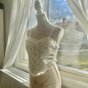 Blommig spetslinne med volangdetaljer - Charmigt vitt linne med blommigt mönster och spetsdetaljer. Linnet har smala axelband och volang längs kanten, vilket ger en romantisk touch. Perfekt för en somrig look! Den är stretchig så passar storlek 34-36, eller XS-S.