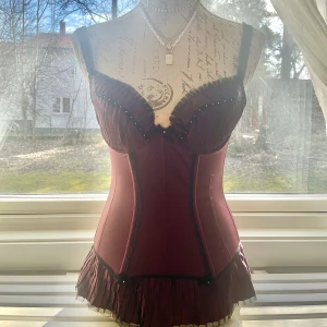 Vinröd korsett med volangdetaljer - Snygg vinröd korsett med svarta axelband och volangdetaljer i nederkant. Den har en elegant design med små pärlor längs bysten och en figurnära passform. Perfekt för att ge en extra touch till din outfit. Är ganska smal men är stretchig och passar de som har lite större byst som storlek 75B och 75C.