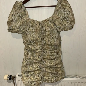 Blommig klänning från NA-KD - Säljer en söt blommig klänning från NA-KD med puffiga kortärmar och rynkade detaljer. Klänningen har en v-ringad design och är perfekt för sommarens alla tillfällen. Den har en härlig mix av gröna och beige toner med små blommor.