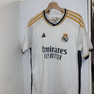 Vit Real Madrid fotbollströja från Adidas - Säljer en vit Real Madrid fotbollströja från Adidas med korta ärmar och guldfärgade detaljer. Tröjan har klubbens emblem på bröstet och sponsorlogga framtill. Perfekt för alla fans som vill visa sitt stöd på matchdagar!