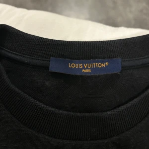 Svart tröja från Louis Vuitton - Säljer en stilren svart tröja från Louis Vuitton med diskret mönster. Tröjan har en liten bröstficka och korta ärmar. Perfekt för en elegant och avslappnad look.