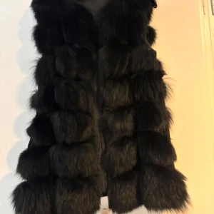 Svart pälsväst äkta - Snygg svart pälsväst med fluffig textur. Perfekt för att hålla sig varm och stilren under kyliga dagar. Västen har en elegant design som passar till många olika outfits.