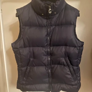 Mörkblå dunväst från Gant storlek M - Säljer en stilren lite mörkt blåaktig dunväst från Gant med dragkedja och hög krage. Perfekt för kyligare dagar och ger en sportig look. Västen har en broderad logga på insidan och är både praktisk och snygg. Storlek M